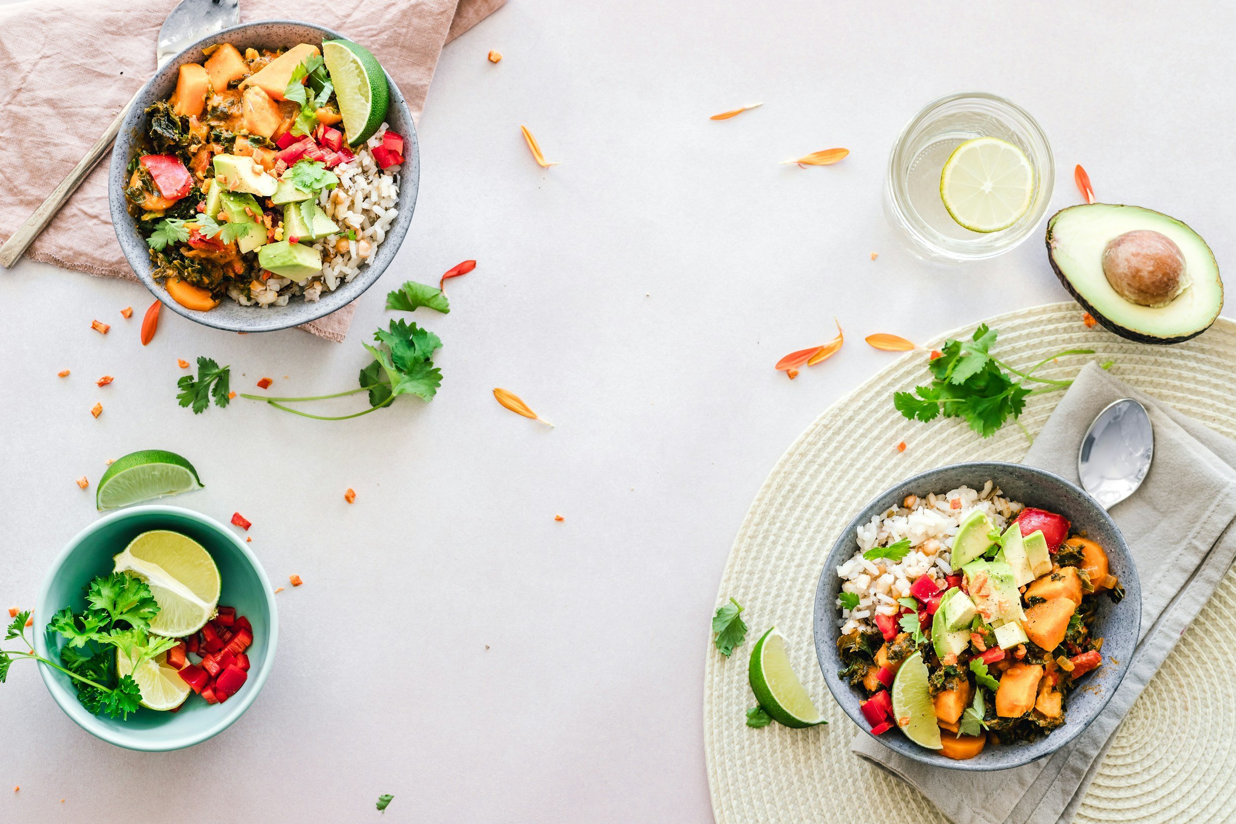 Bowls of vegetarian rice and vegetable bowls garnished with lime wedges, avocado slices, and fresh cilantro on a light-colored table with scattered flower petals and small bowls of sliced lime, cilantro, and red peppers.