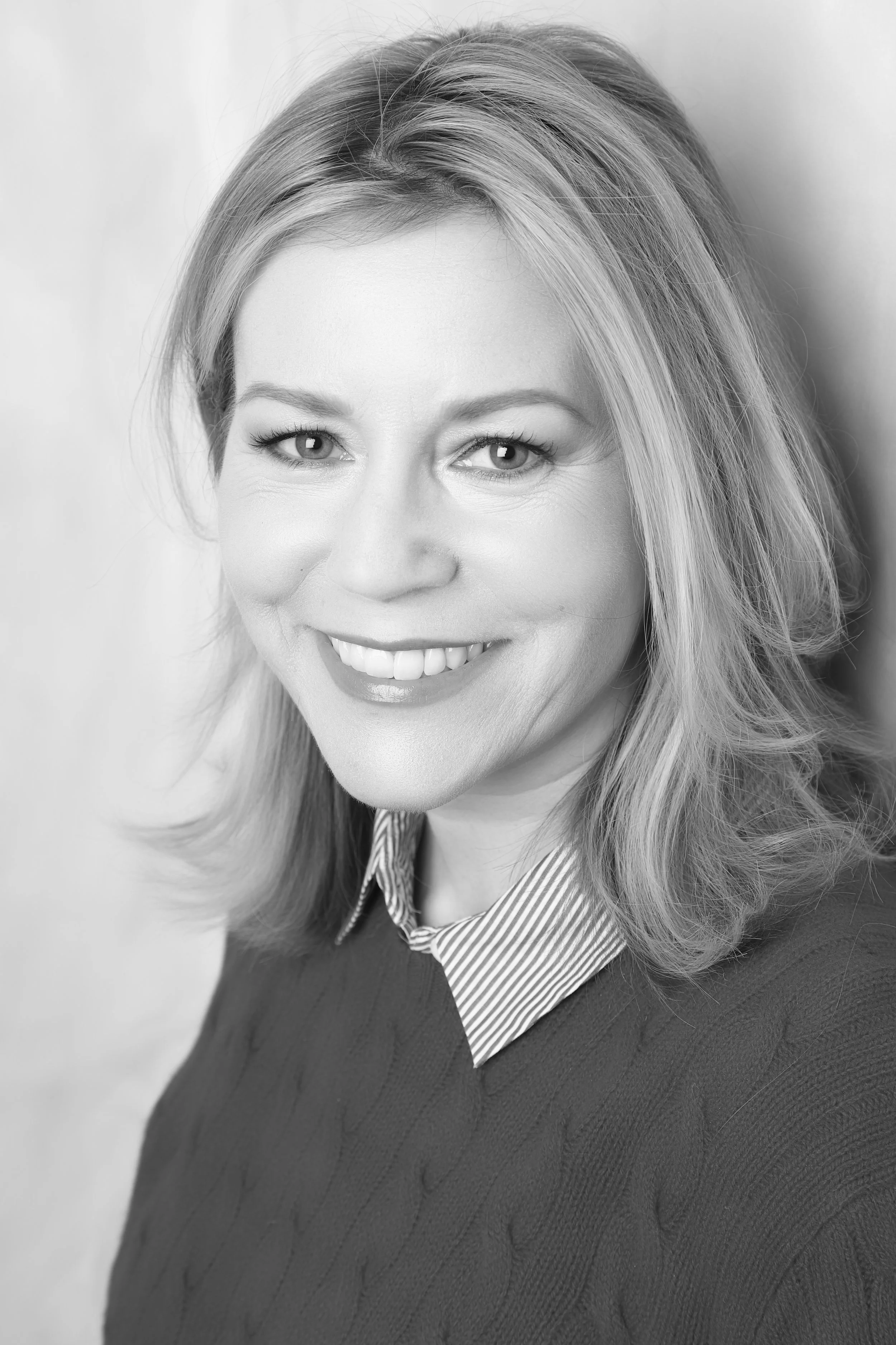 Close-up black and white photo of a woman with shoulder-length hair, smiling, wearing a collared shirt and a sweater.