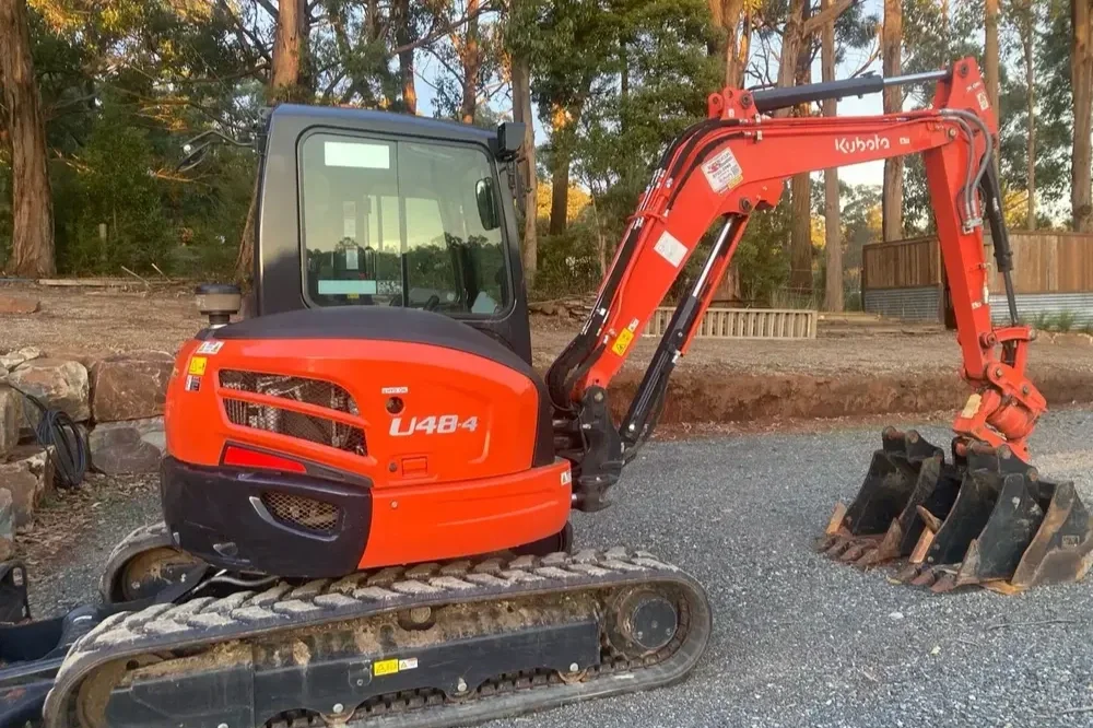 Orange Kubota mini excavator with black tracks and a bucket attachment, parked outdoors on gravel, with trees and a building in the background.