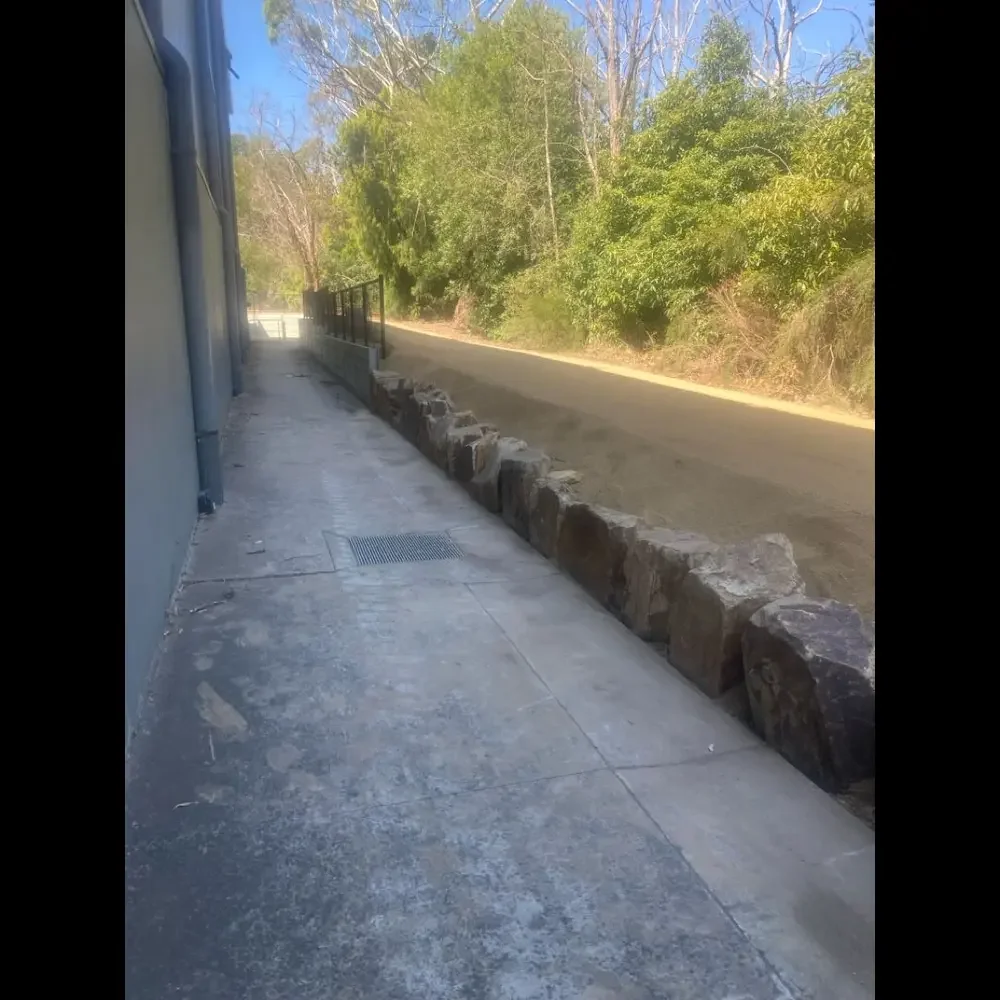 Sidewalk with a row of large rocks acting as a barrier separating it from a dirt road, with trees and bushes in the background under a clear blue sky.