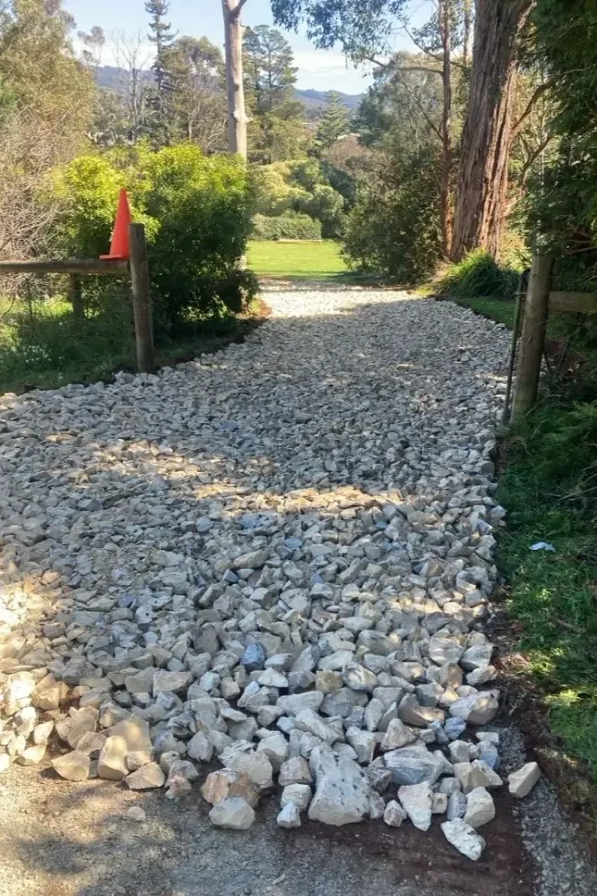 Gravel pathway lined with trees and bushes, with a wooden fence and orange construction cone on the left side.