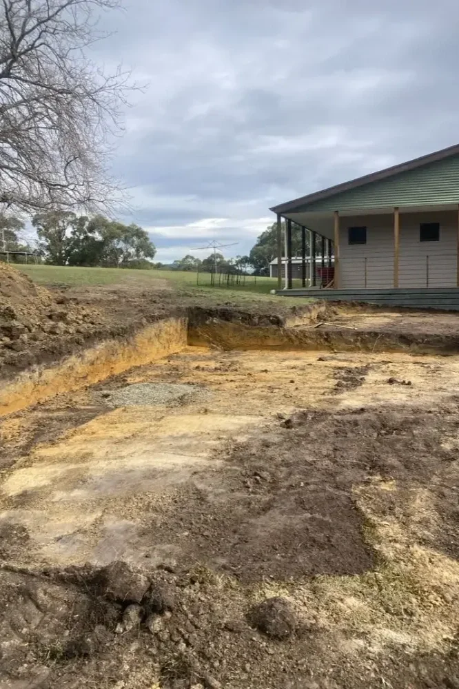Bare ground and soil excavation site near a house with a porch and green lawn under cloudy sky.