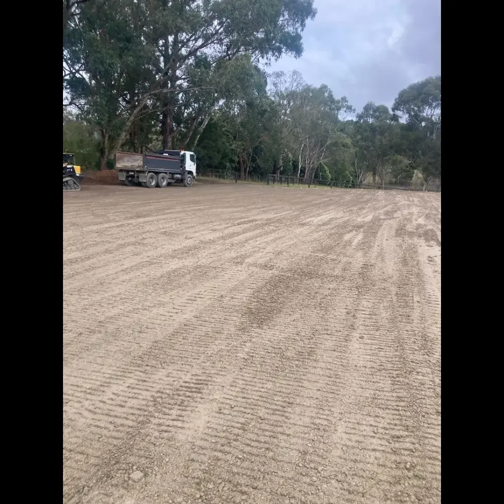 A large, cleared outdoor area with freshly graded dirt, two construction vehicles near the trees, and a background of trees and overcast sky.
