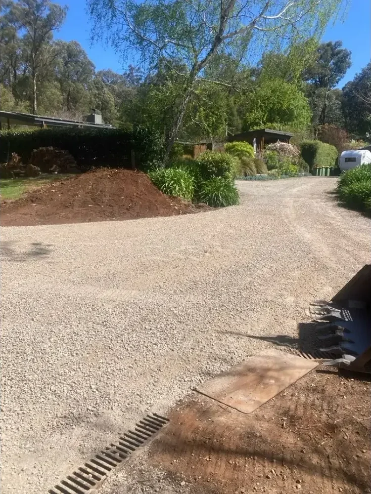 A gravel driveway in a residential area with trees, bushes, a house, and a small trailer or shed in the background.