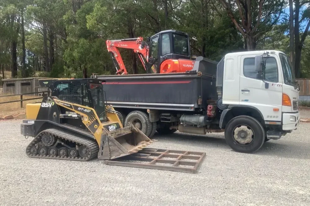A construction site with a white truck, an orange excavator in the truck bed, and a small yellow tracked bulldozer in front of the truck on gravel ground.