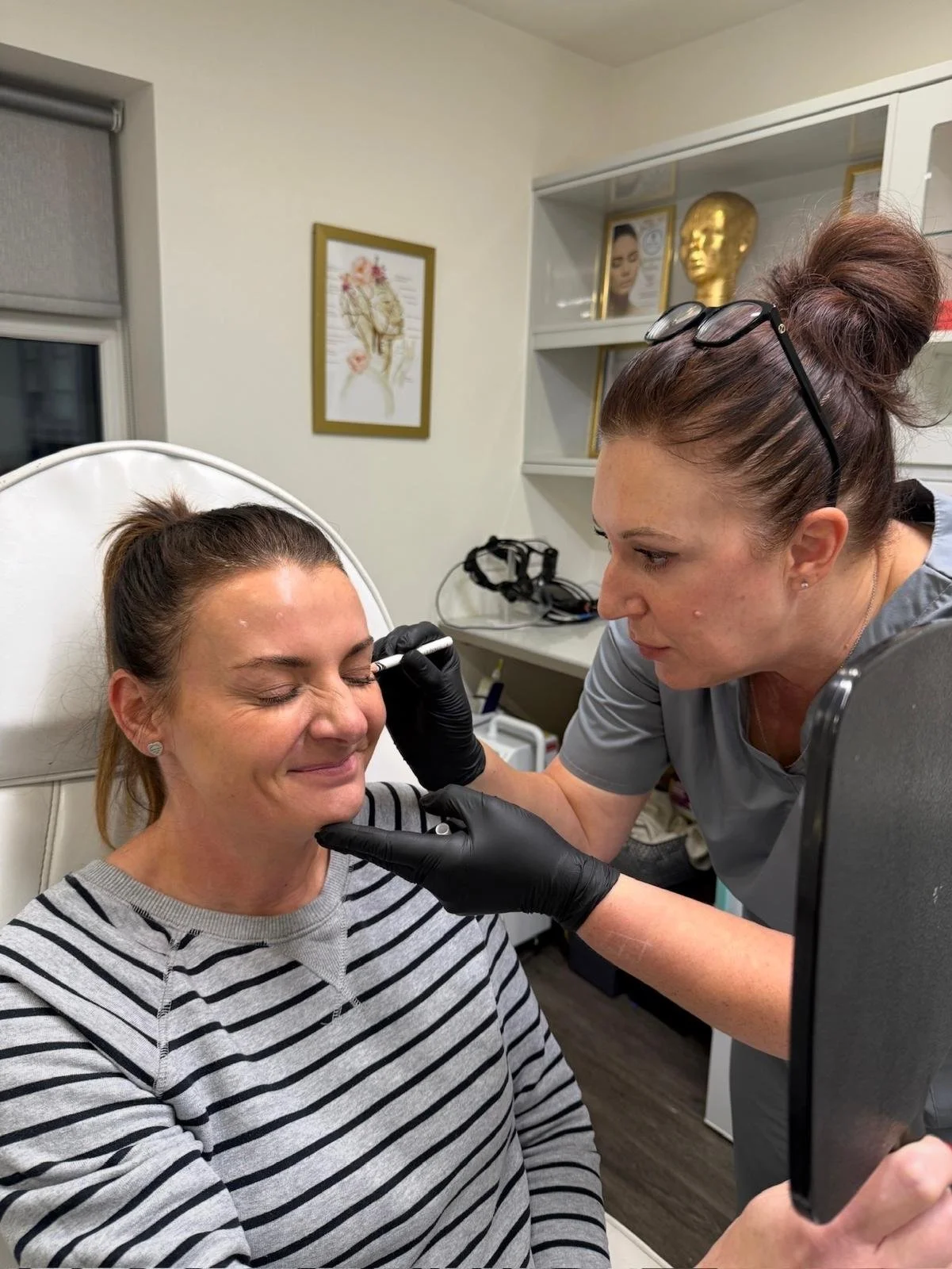 A woman in striped shirt sitting in a medical chair with her eyes closed, receiving a procedure from a healthcare professional with glasses on her head and black gloves, in a medical or skincare clinic.