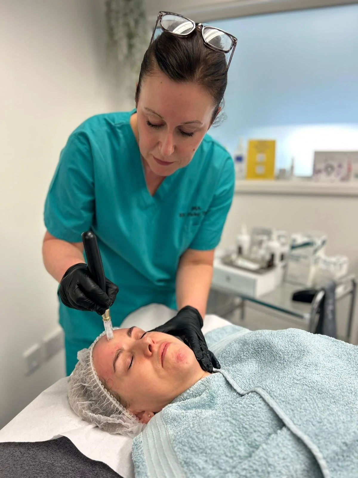 A woman with glasses on her head and wearing teal scrubs administers a cosmetic facial treatment with a syringe to a woman's face in a clinical setting.