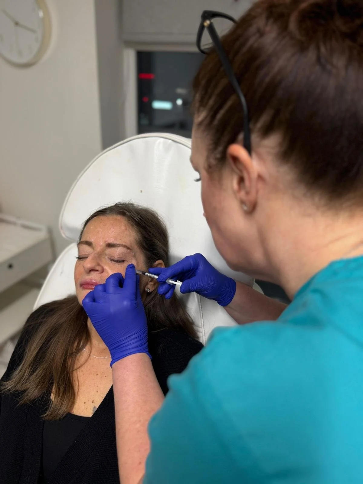 A woman receiving a cosmetic treatment in a medical setting, lying back in a chair with eyes closed, as a healthcare professional with glasses and blue gloves administers an injection to her face.