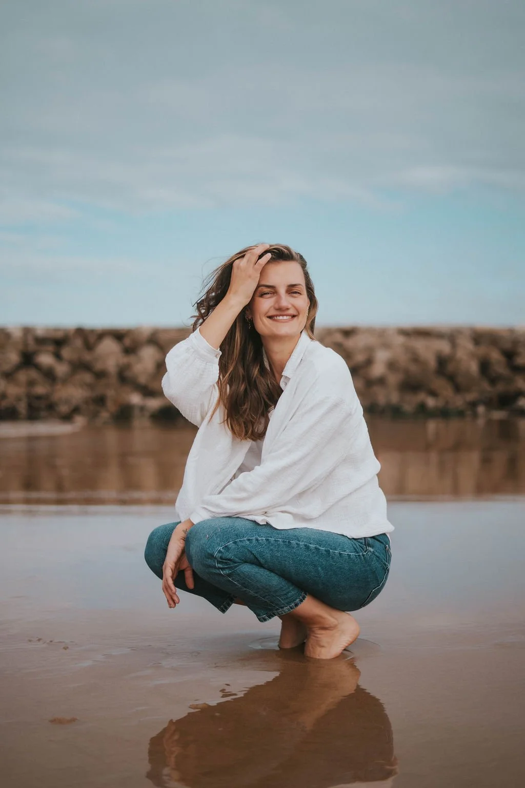 Woman smiling, squatting in shallow water at the beach, with rocks and sky in the background.