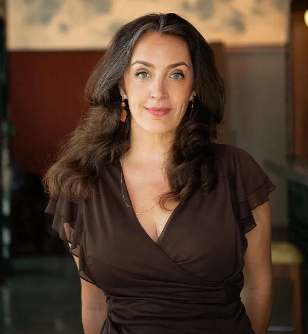 Portrait of a woman with long, wavy brown hair, wearing a dark brown dress with ruffled sleeves, gold earrings, and a delicate necklace, standing in an indoor setting with warm lighting and blurred background.