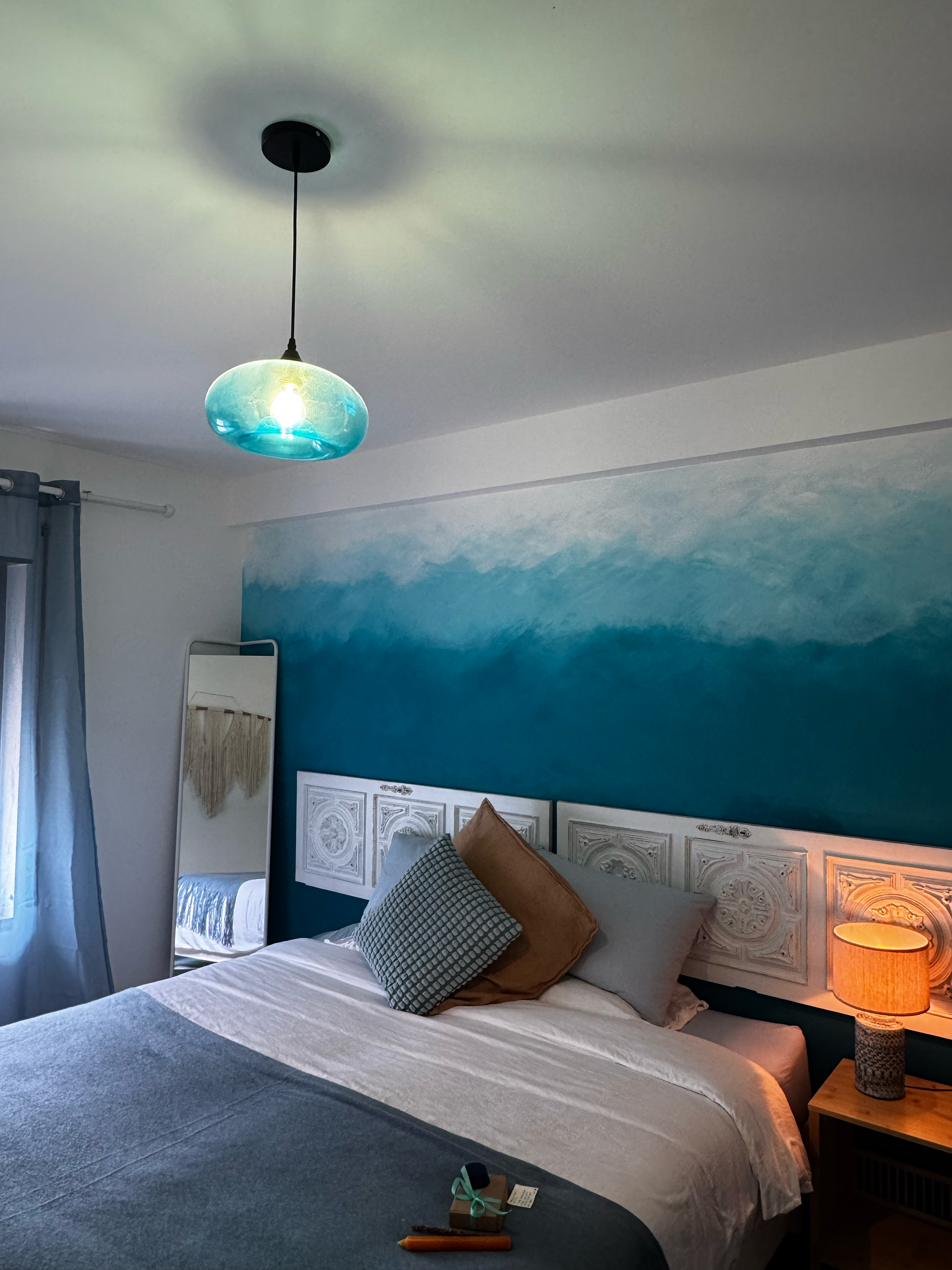 A bedroom with a wall mural of a cloudy sky in blue and white, a bed with pillows, a nightstand with a lamp, a mirror, and a ceiling light with a blue glass shade.