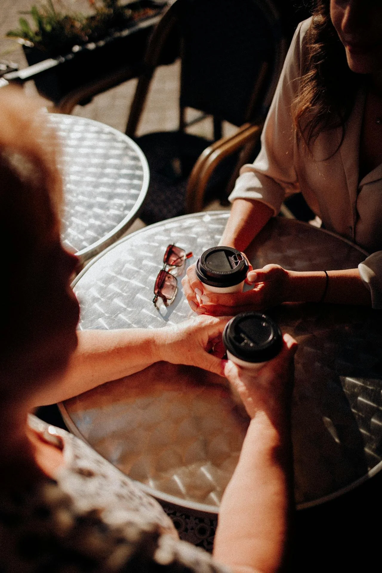 Two women sitting at a metal table, holding coffee cups in a cozy setting, with glasses, a beige blouse, and a leopard print top visible.