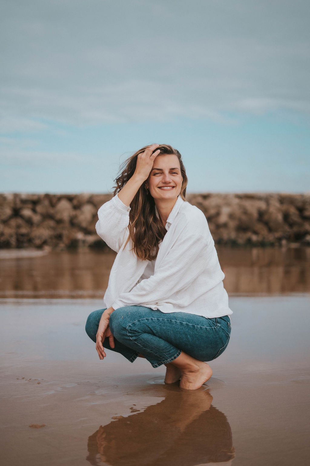 A woman crouches knee-deep in shallow water, smiling, with her hand in her hair, at the beach with rocks and cloudy sky in the background.