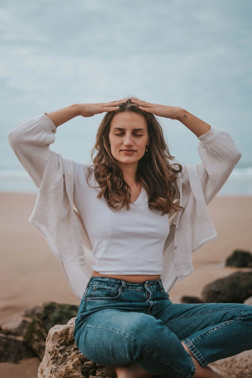 A woman sits on a rock at the beach with her eyes closed, hands on her head, wearing a white crop top, a light-colored open shirt, and blue jeans, with the ocean and cloudy sky in the background.
