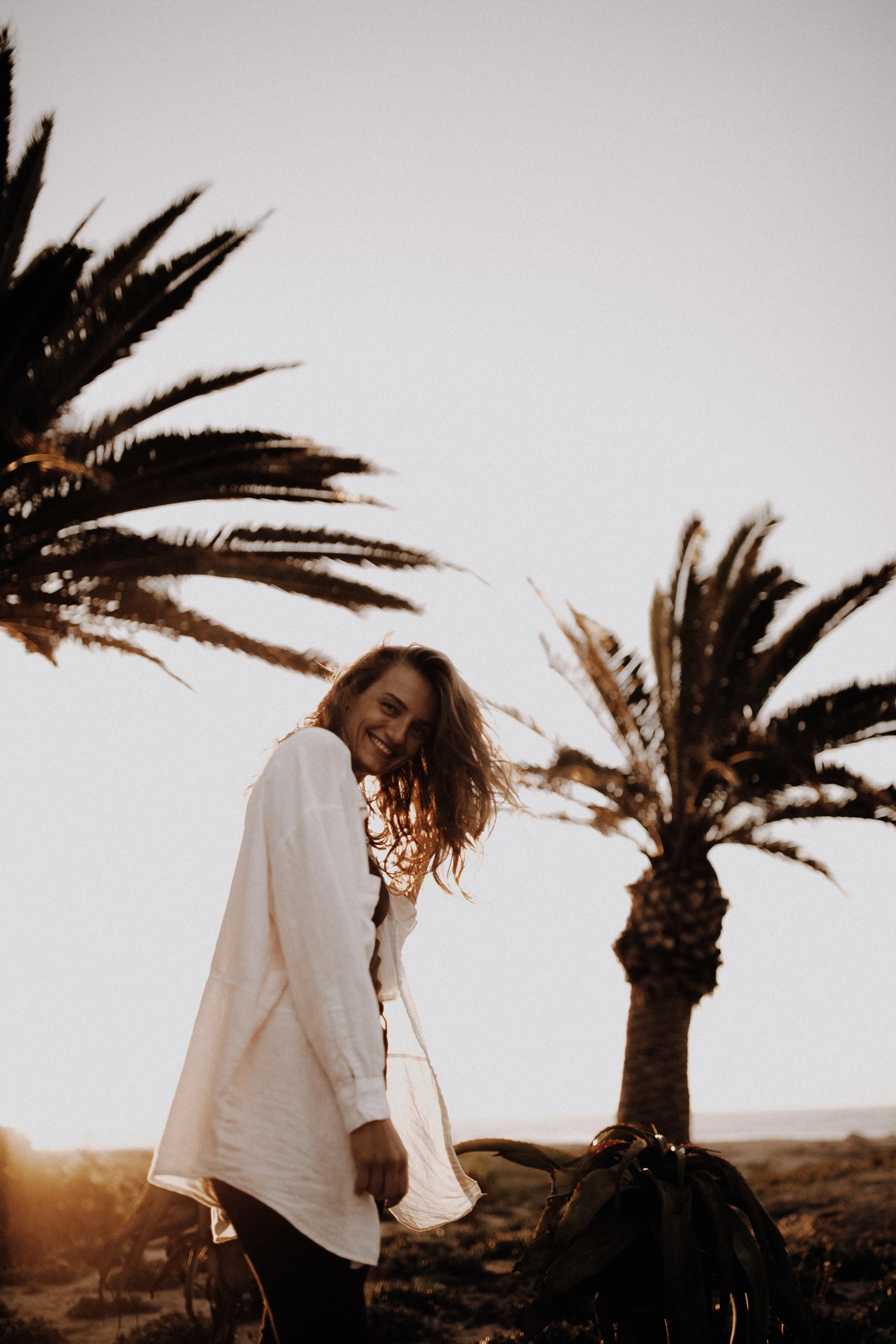 A smiling woman with curly hair standing outdoors near palm trees during sunset, wearing a white jacket over a black top.