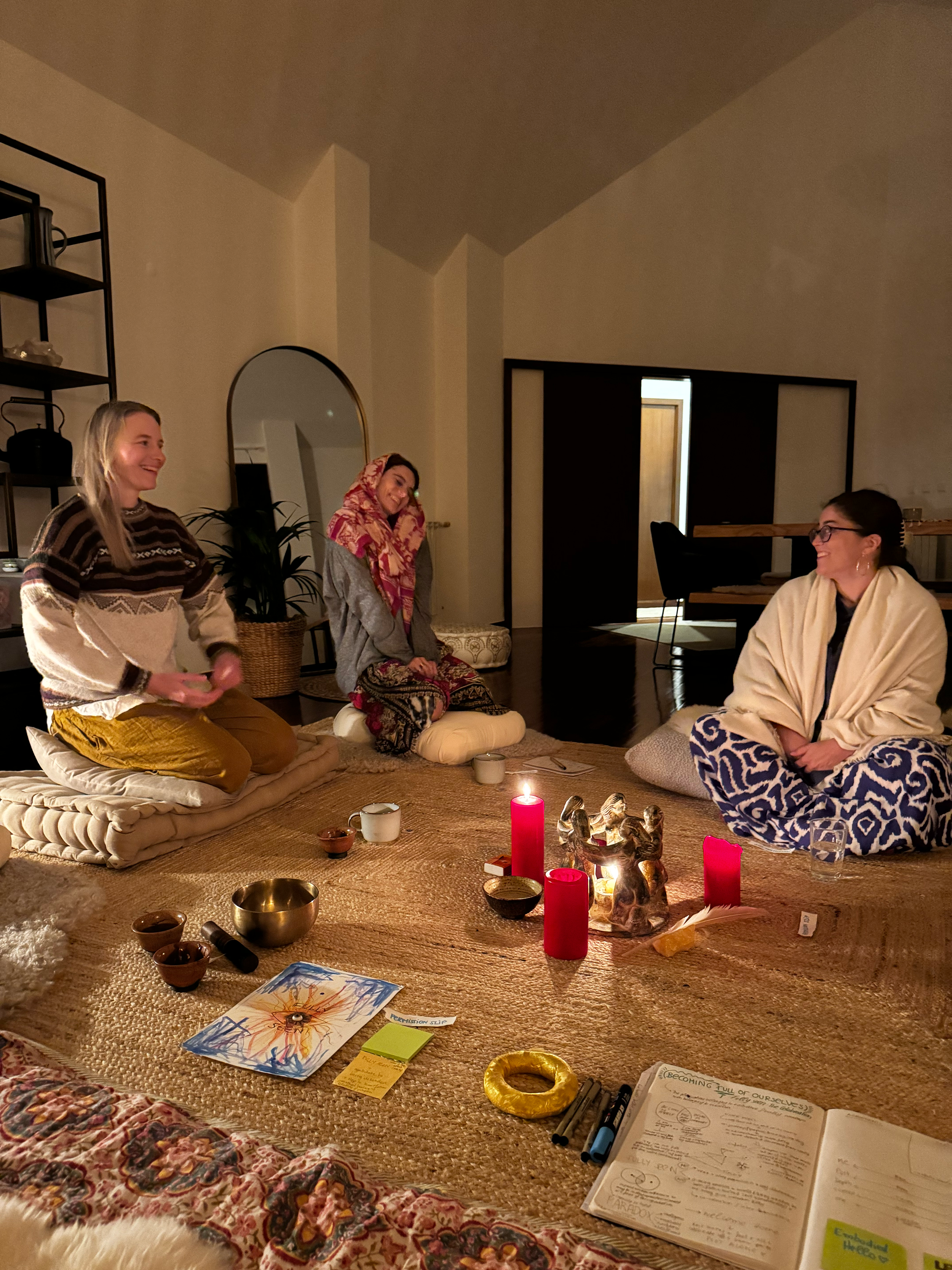 Three women sitting on a carpeted floor in a dimly lit room, engaging in a spiritual or meditation ritual with candles, statues, and writing materials on the floor.