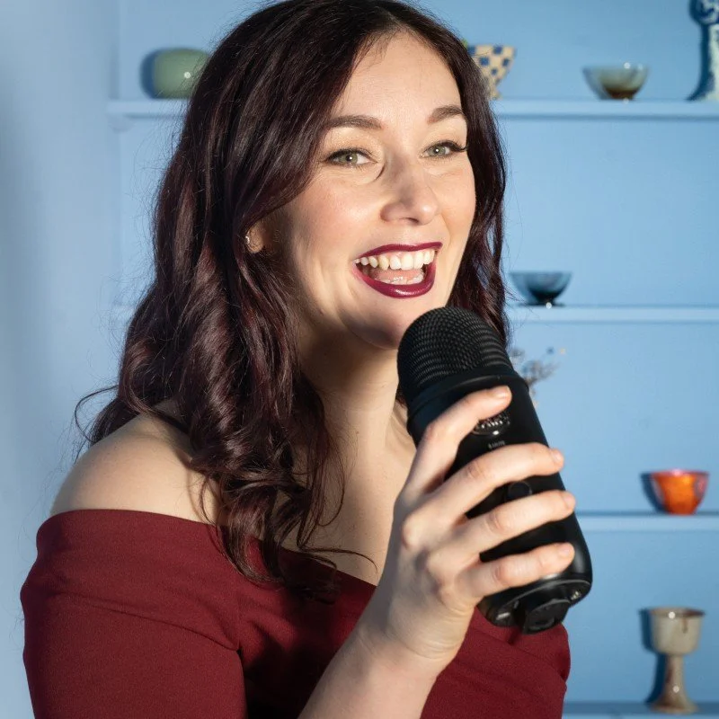 Woman with dark red hair, wearing an off-the-shoulder maroon top, smiling and holding a microphone.