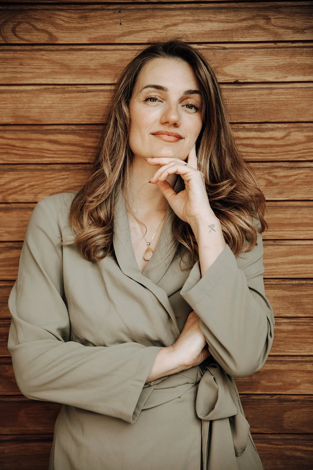 A woman with long, wavy brown hair standing against a wooden wall. She is wearing a beige jacket, has her right hand resting under her chin, and is smiling slightly.