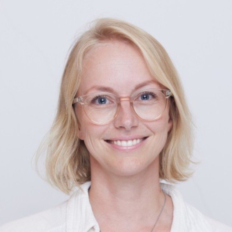 A woman with blonde hair and glasses smiling at the camera against a light gray background.