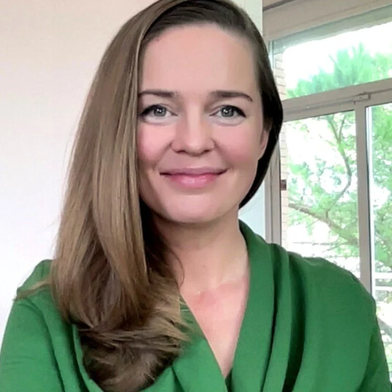 A woman with long brown hair and blue eyes smiling, wearing a green top, standing indoors in front of a window with trees outside.