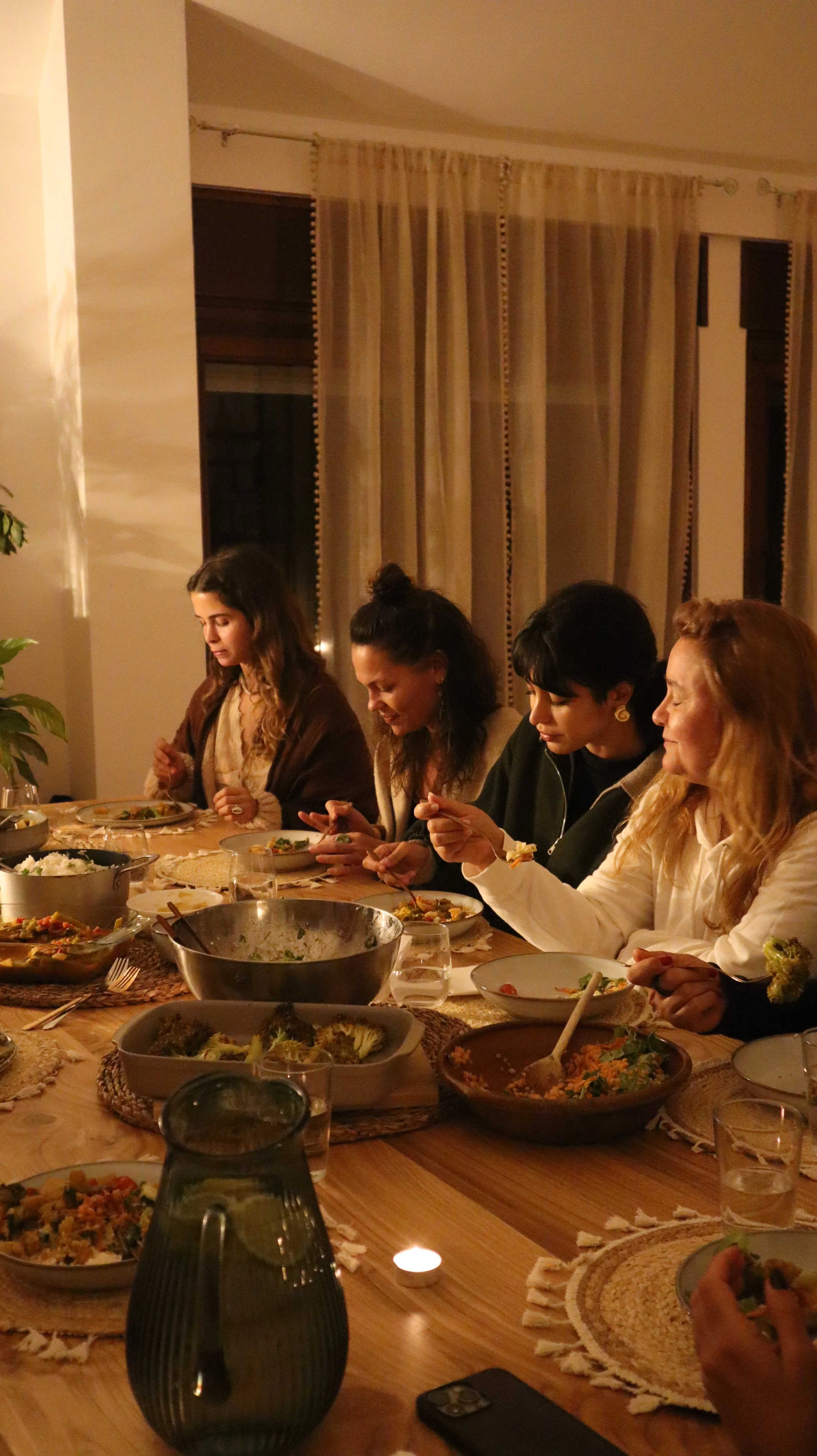 A group of women sitting at a dinner table enjoying a meal together, with various dishes and drinks on the table in a warmly lit room.