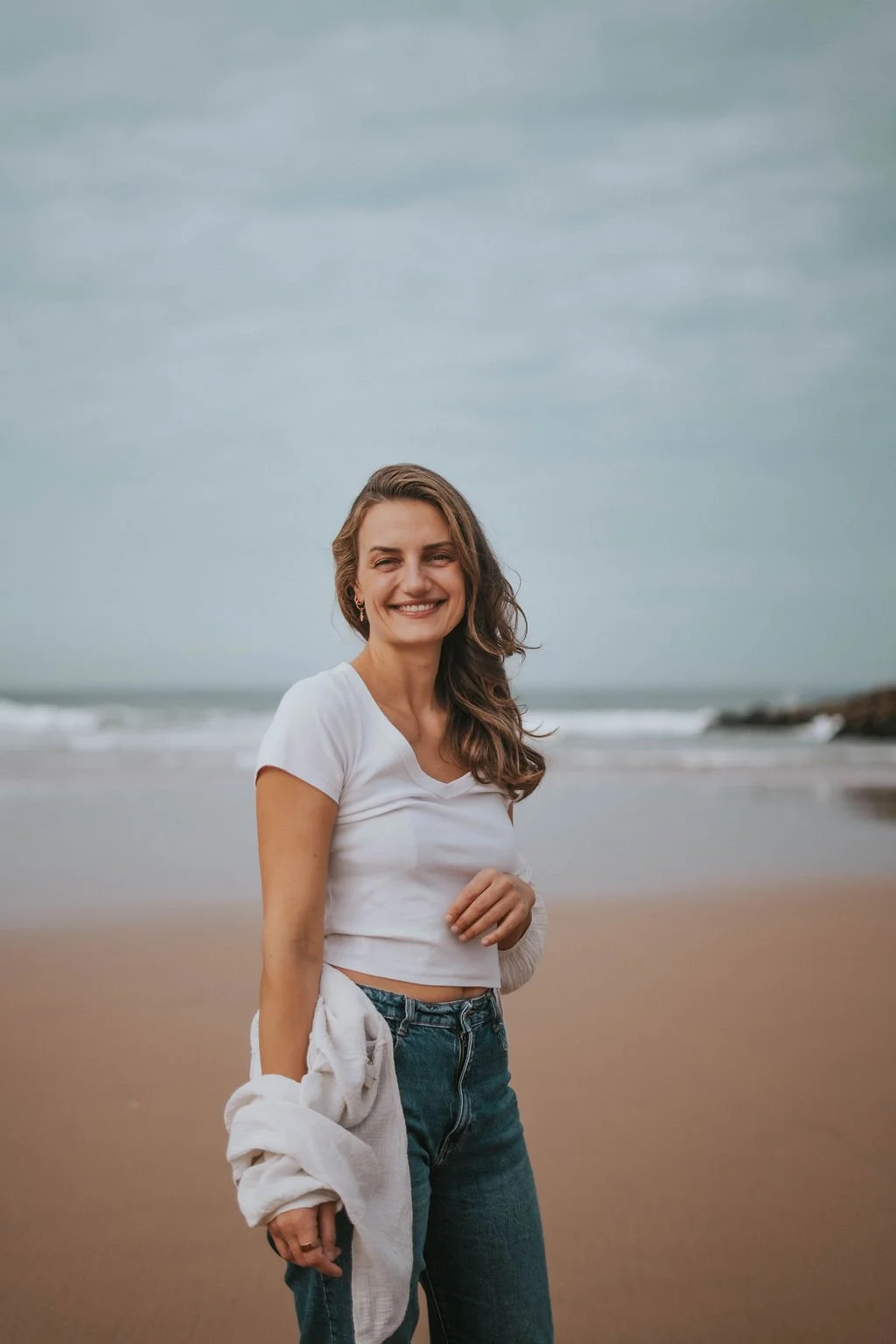 A woman smiling at the beach, holding a white towel, wearing a white t-shirt and jeans, with ocean waves and a cloudy sky in the background.