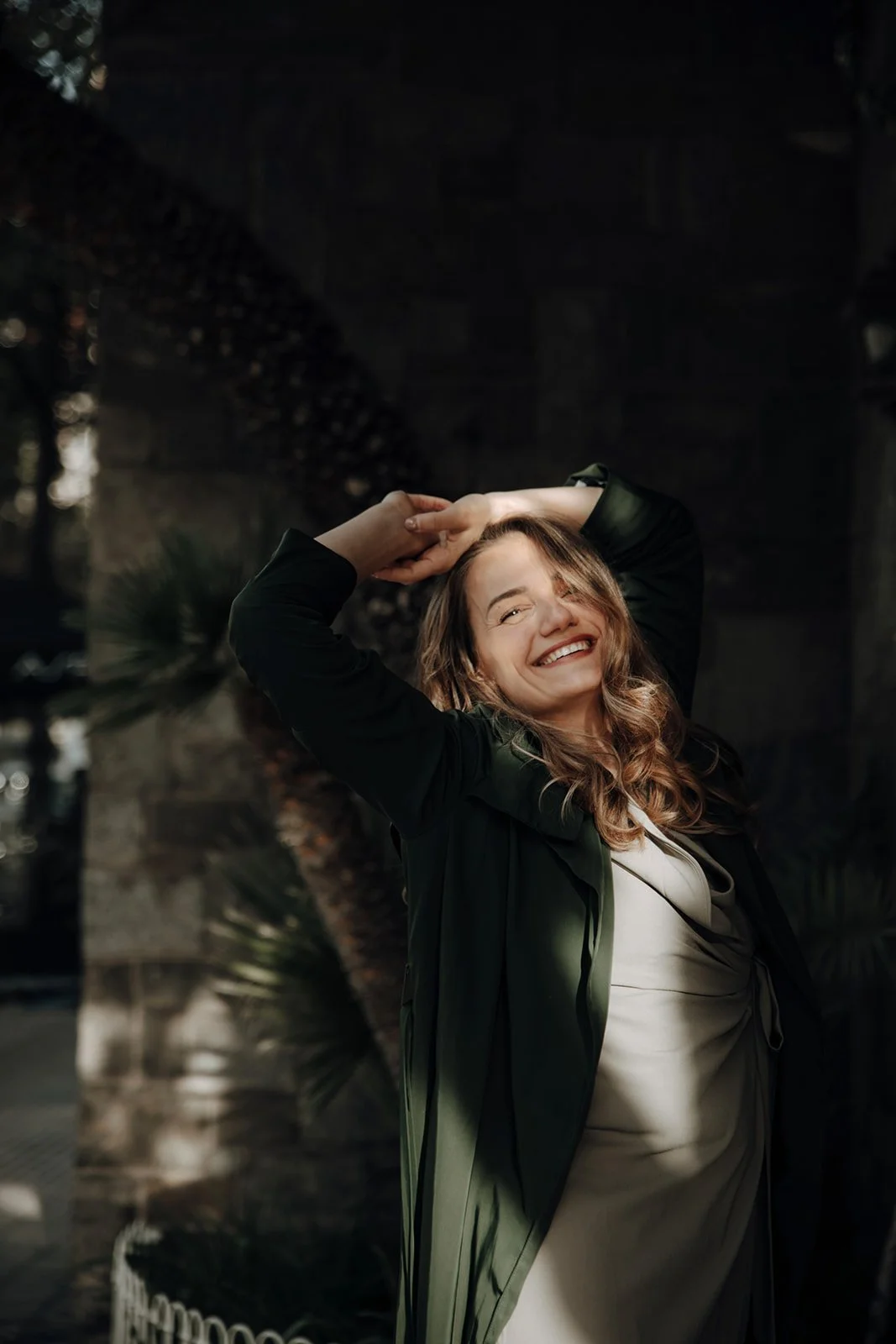 A smiling woman with long, wavy hair posing outdoors at sunset, wearing a light beige dress and a dark green jacket, with her hands raised above her head.