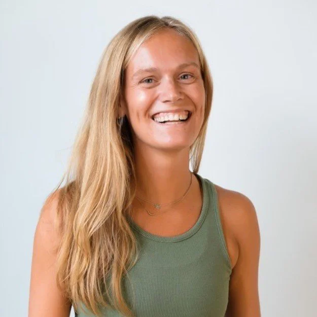Young woman with long blonde hair, smiling, wearing a green sleeveless top, against a plain light background.