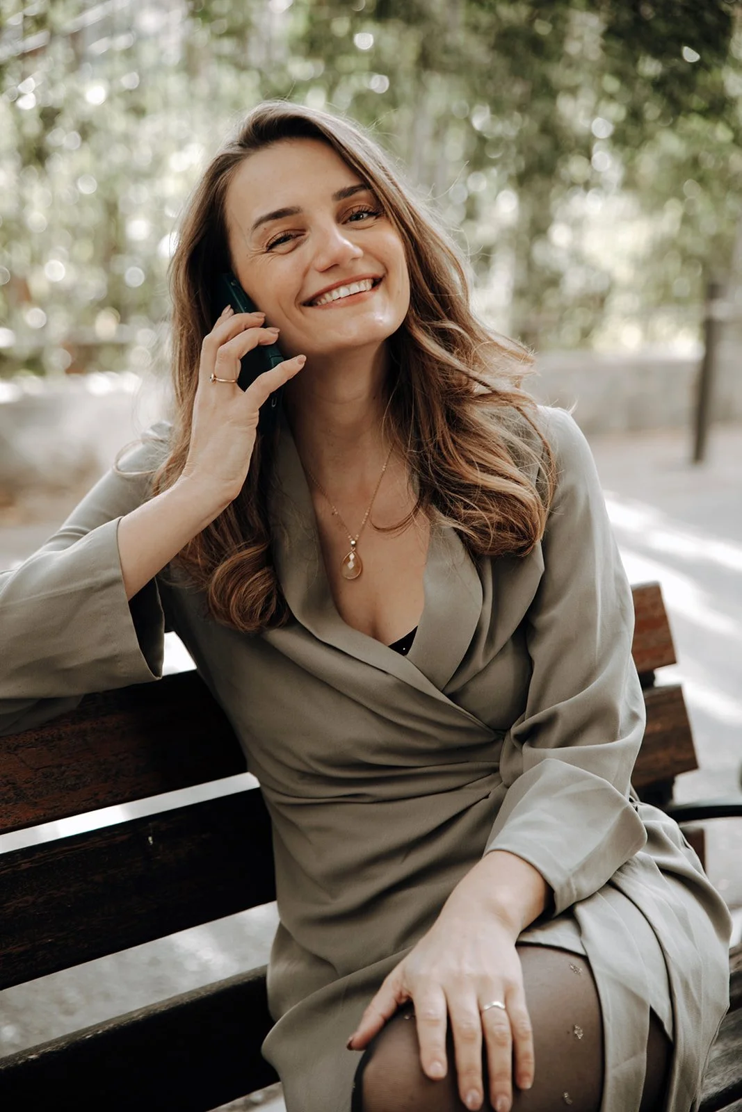 A woman with long wavy hair, smiling while talking on a phone, sitting on a wooden bench outdoors with a blurred background of trees.