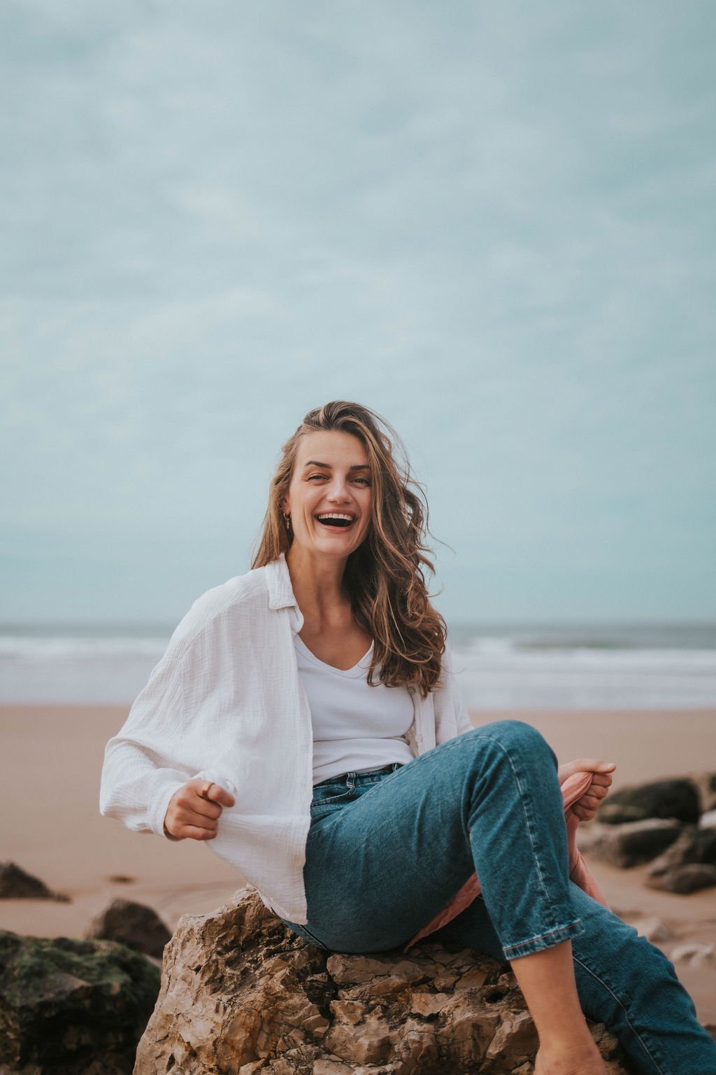 A woman sitting on a rock at the beach, smiling and looking happy, with sand, rocks, and the ocean in the background.