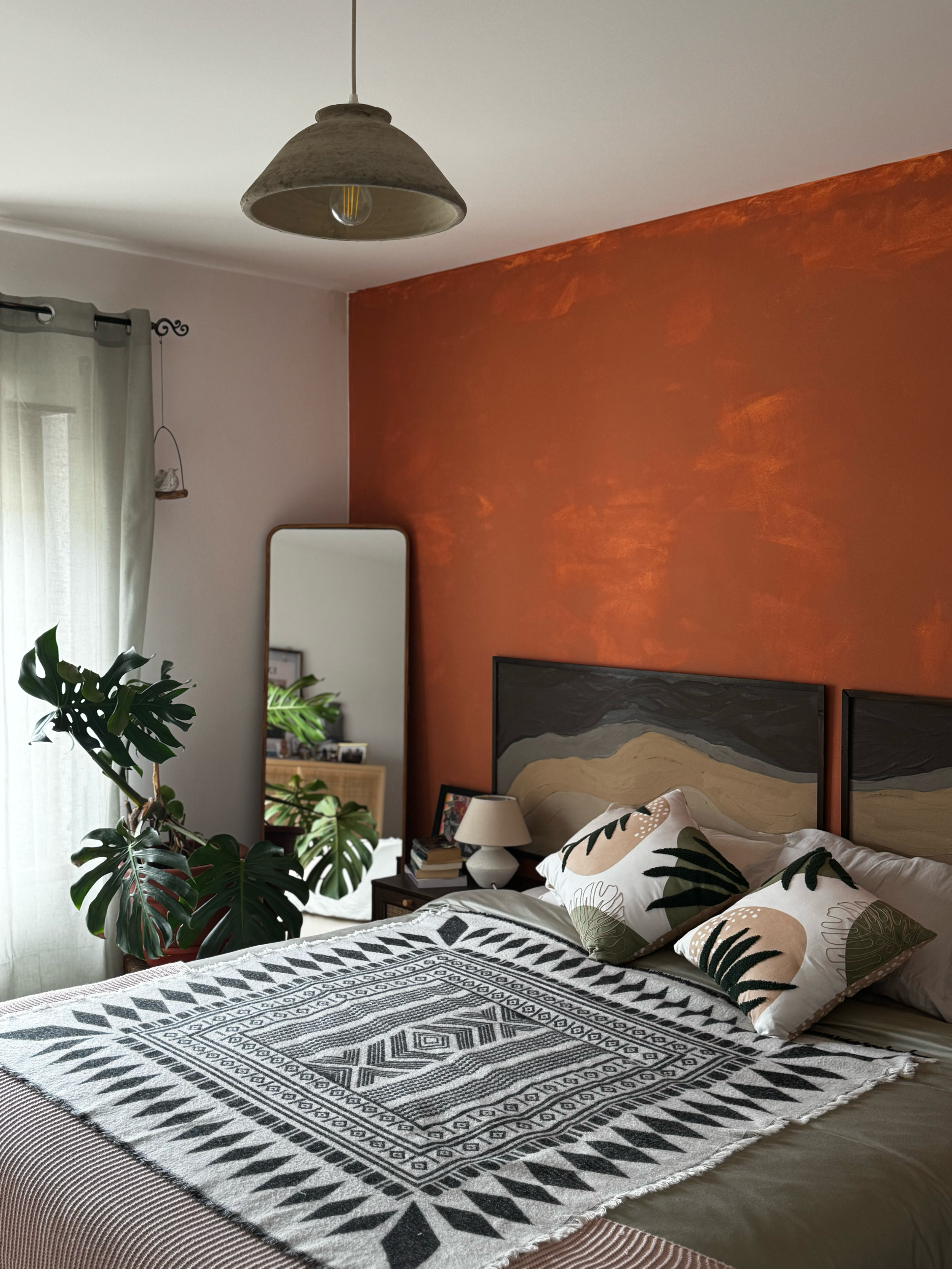A cozy bedroom with a bed, decorative pillows, a black and white geometric patterned throw blanket, a nightstand with a lamp, a mirror, and a large houseplant near the window, with an orange accent wall.
