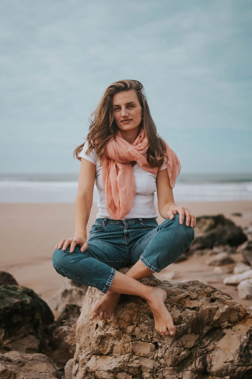 A woman sitting on a large rock at the beach, with ocean waves and a cloudy sky in the background.