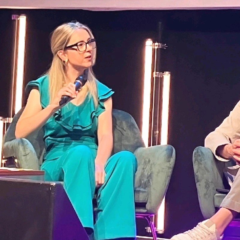 A woman with long blonde hair and glasses is sitting on a green velvet armchair, holding a microphone, wearing a turquoise dress. Another person is partially visible on her right, sitting with legs crossed, wearing tan pants and beige sneakers. There are vertical light fixtures behind them.