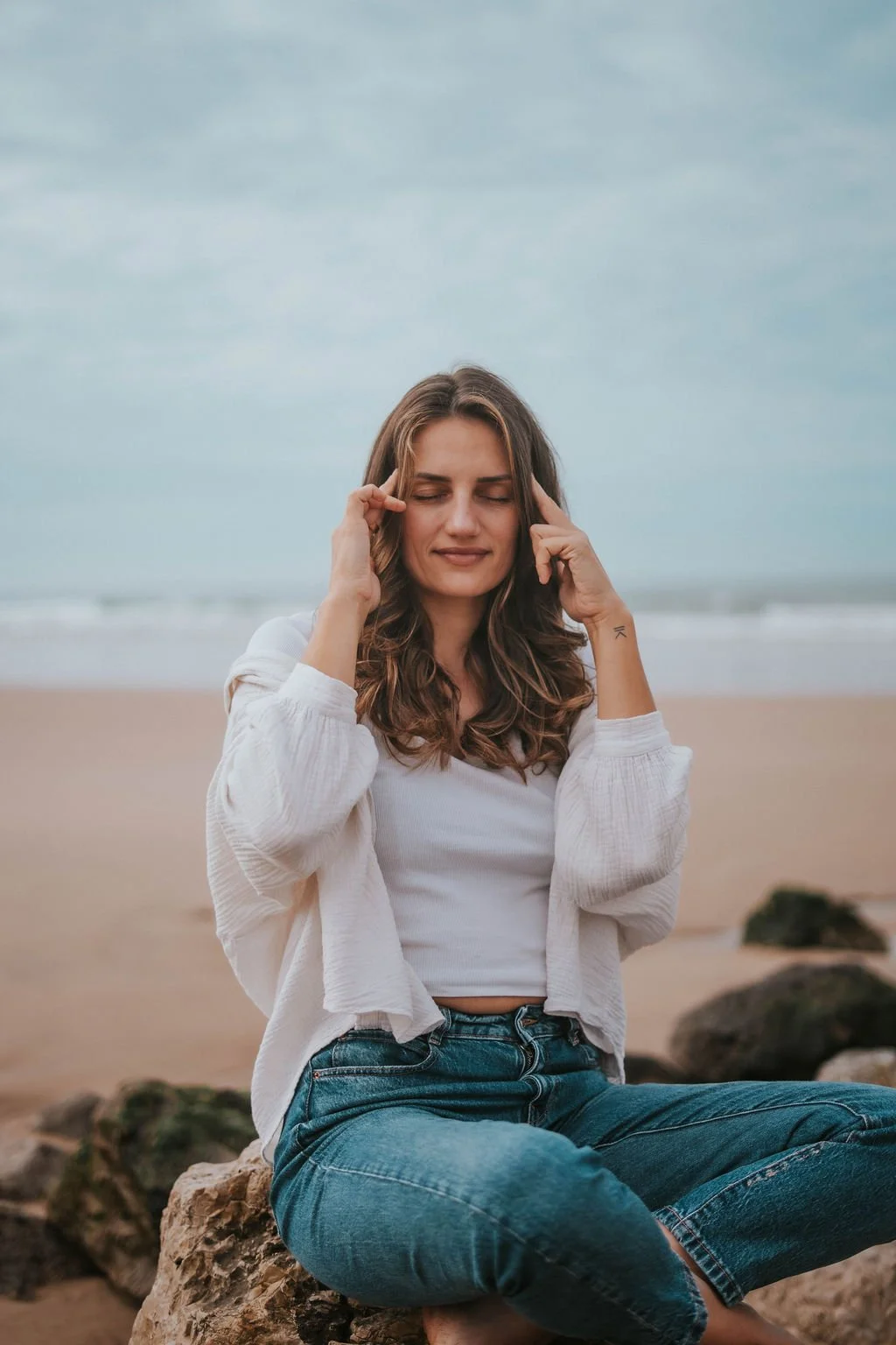 A young woman sitting on rocks at the beach, with eyes closed, holding her head with both hands and smiling softly.