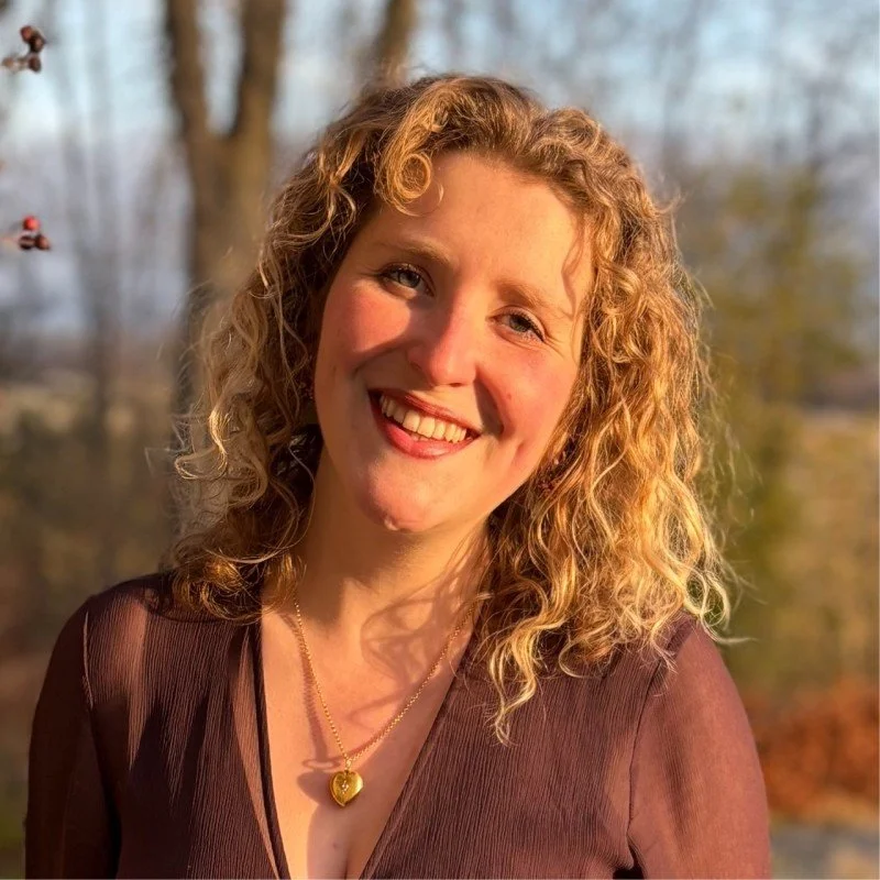 A woman with curly blonde hair wearing a brown top and gold necklace smiling outdoors with trees in the background.