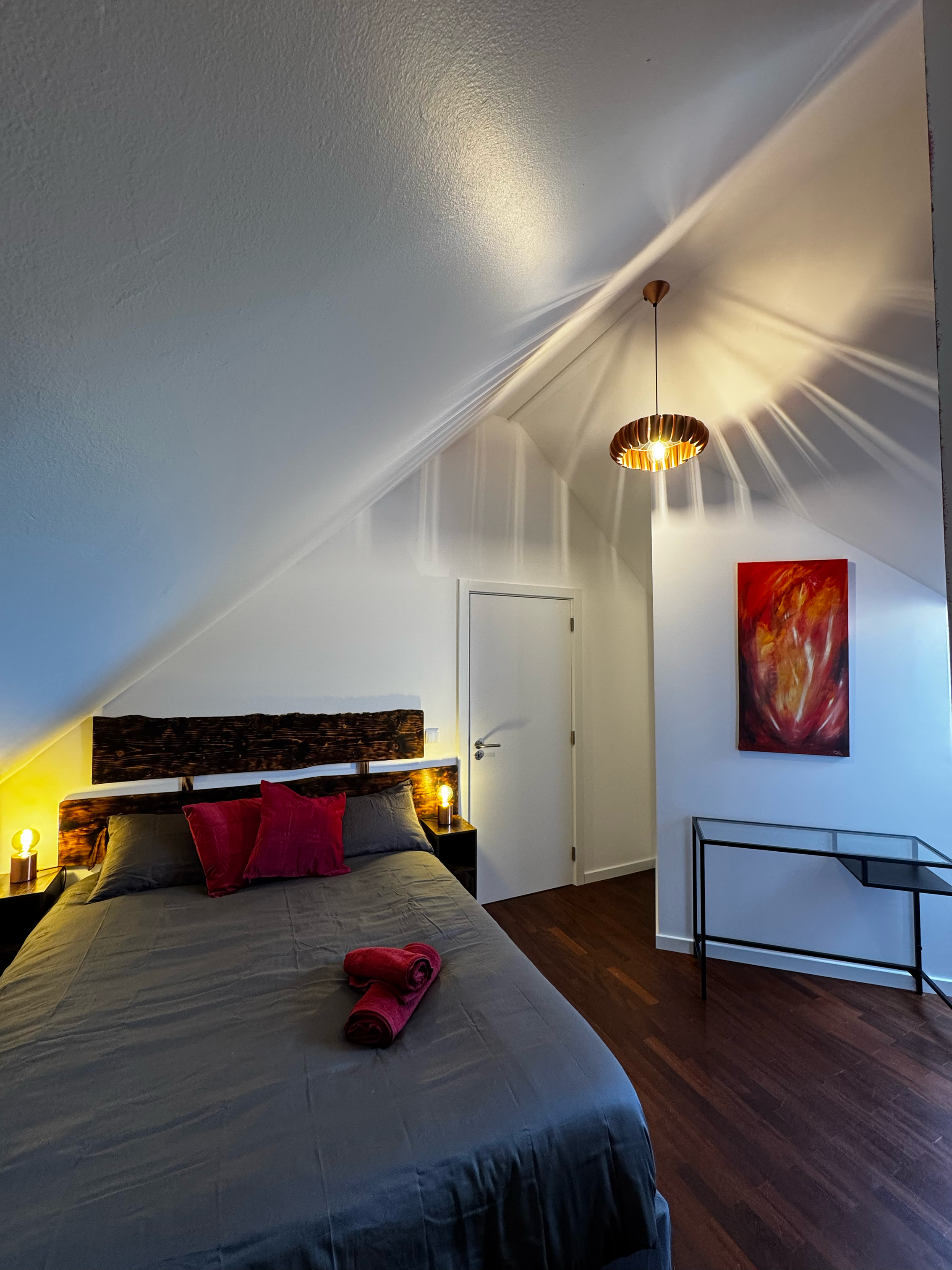 A bedroom with a sloped ceiling, a bed with gray bedding and pink pillows, two bedside lamps, a hanging light fixture, a colorful abstract painting on the wall, and a small glass table.