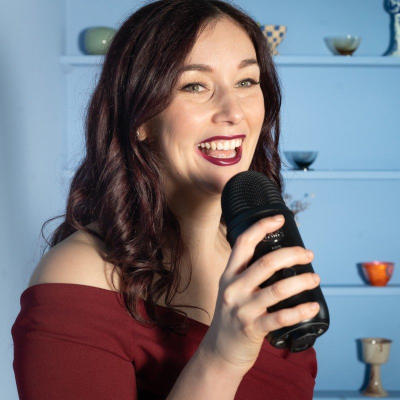 A woman with long dark hair smiling and holding a black microphone, wearing an off-the-shoulder red top, in front of a blue background with shelves and colorful bowls.