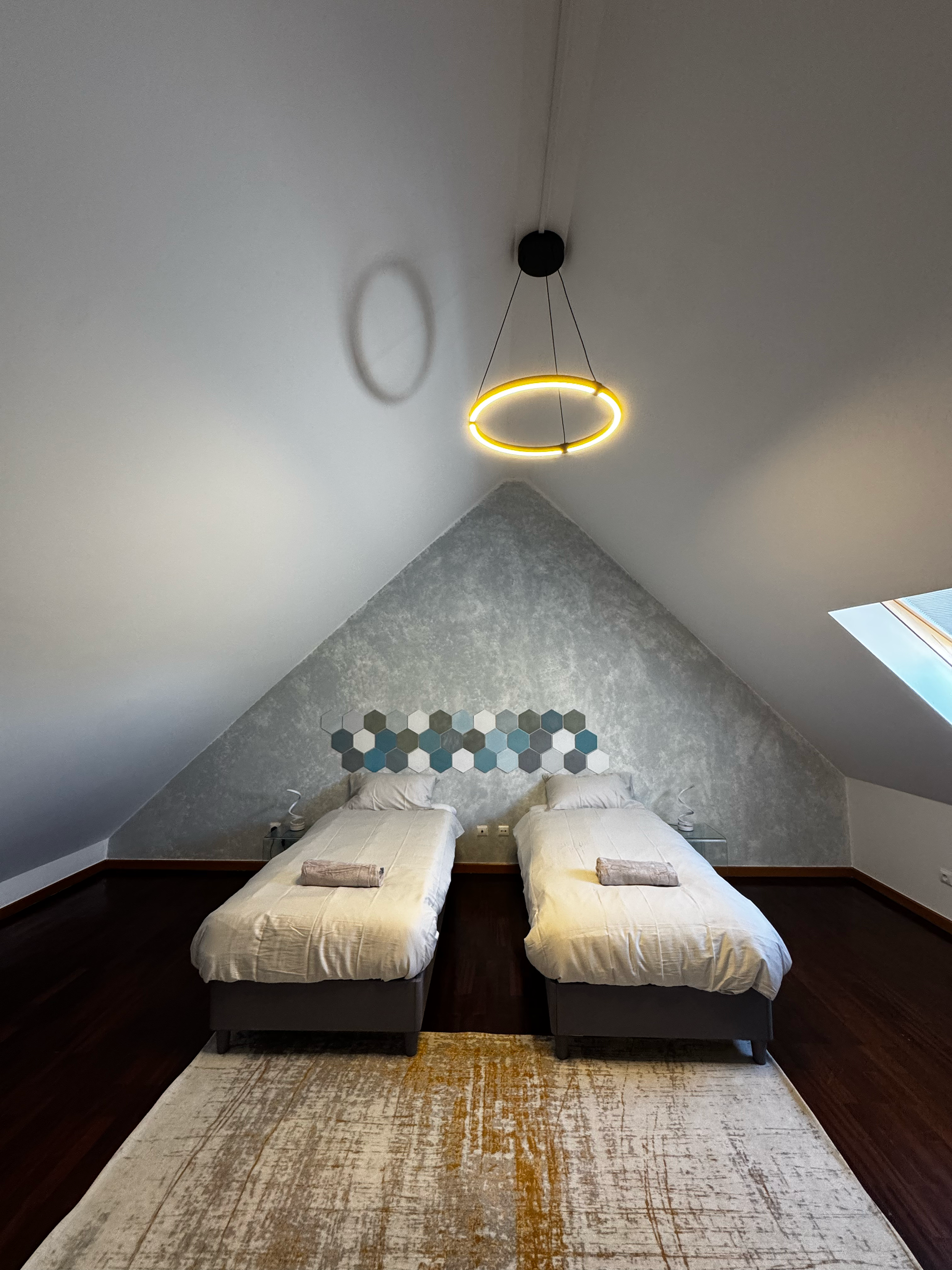 Two single beds in an attic bedroom with a sloped ceiling, grey and white wall decor, a pendant circular light fixture, and a small skylight window.
