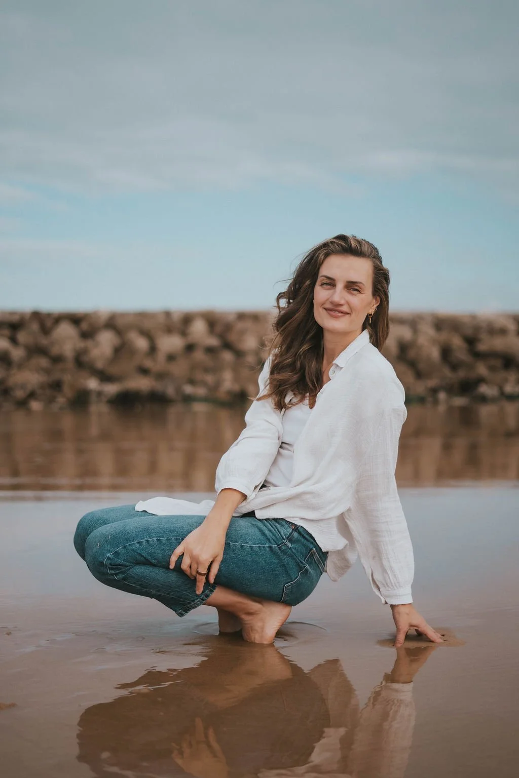 A woman with brown hair wearing a white shirt and blue jeans squatting in shallow water near the beach, smiling with her hand touching the water, in front of a rocky shoreline and cloudy sky.