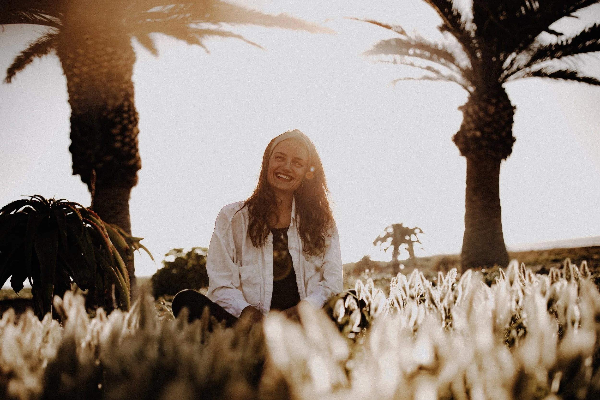 A woman sitting on the ground among white succulents, smiling, with palm trees in the background during sunset.