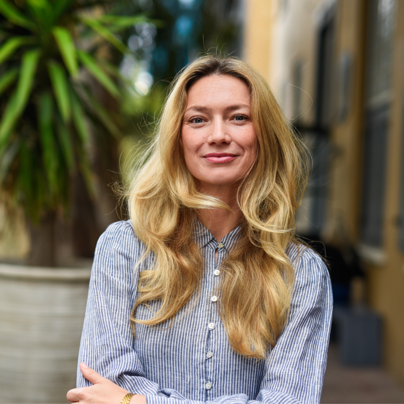 A woman with long, wavy blonde hair wearing a blue and white striped button-up shirt standing outdoors in front of a large potted plant and a yellow building.