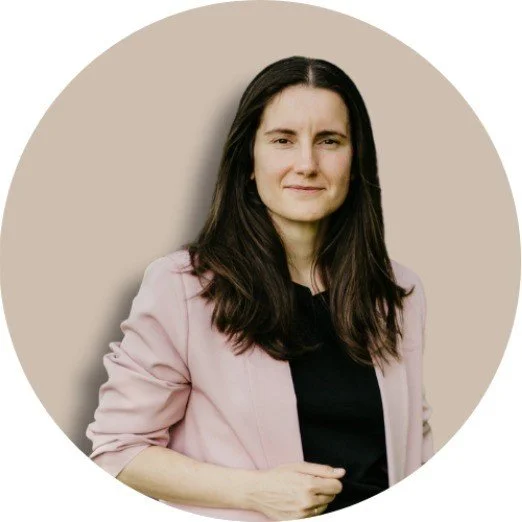 A woman with long dark hair wearing a pink blazer over a black top, standing against a light beige background.