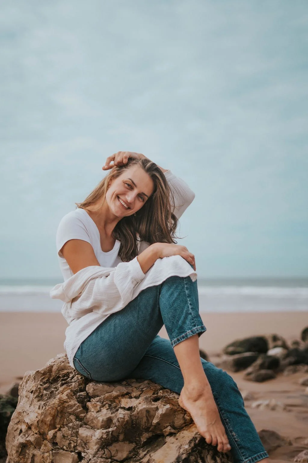 A woman sitting on a large rock at the beach, smiling and looking at the camera, with a cloudy sky in the background.