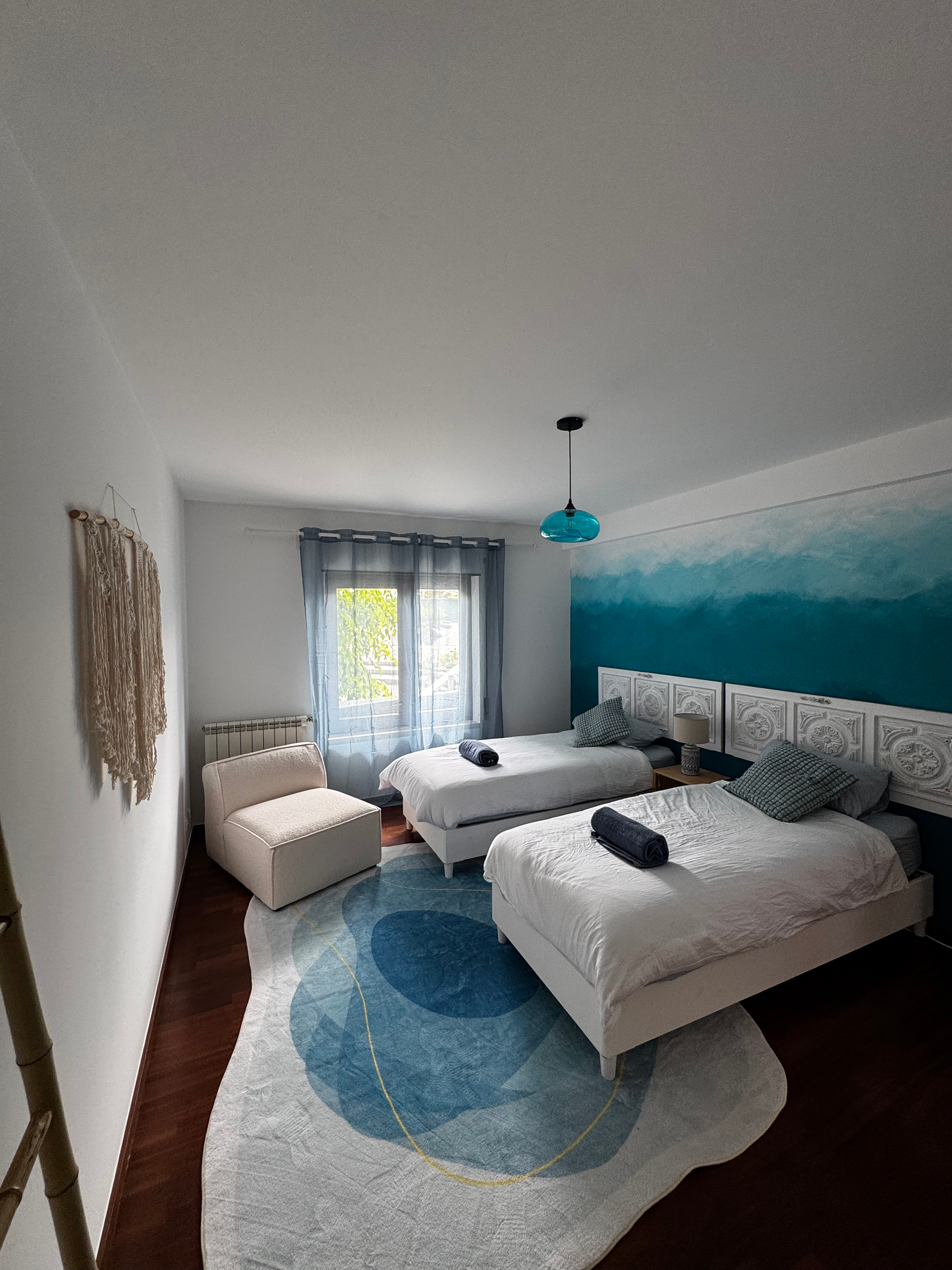 A bedroom with two beds, a white armchair, a colorful abstract rug, a window with sheer curtains, and a blue ocean wave mural on the wall behind the beds.