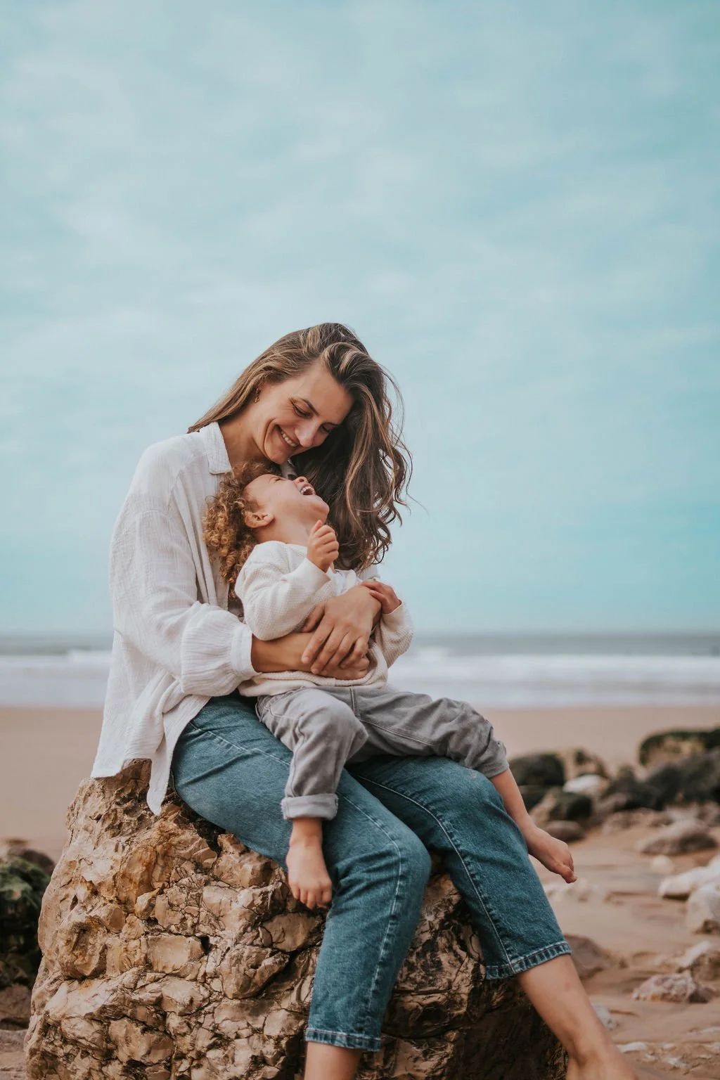 A woman and a young girl sitting on a large rock at the beach, laughing and sharing a joyful moment together. The woman has long brown hair, and the girl has curly hair. Both are wearing white tops, and the background features a sandy beach and the ocean under a cloudy sky.