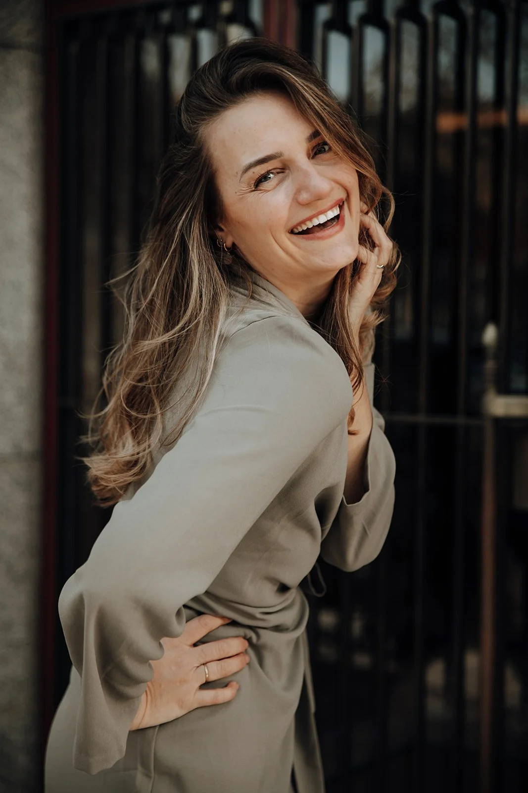 A woman with light brown hair smiling and standing outdoors, wearing a beige blazer, leaning against a black fence with a stone wall to her left.