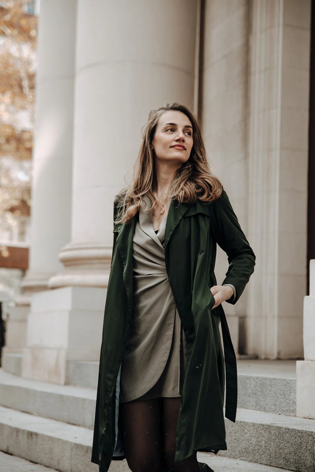 A woman with long, wavy hair standing on steps in front of a building with large columns, wearing a beige dress, black stockings with small embellishments, and a dark green trench coat, looking off to the side.