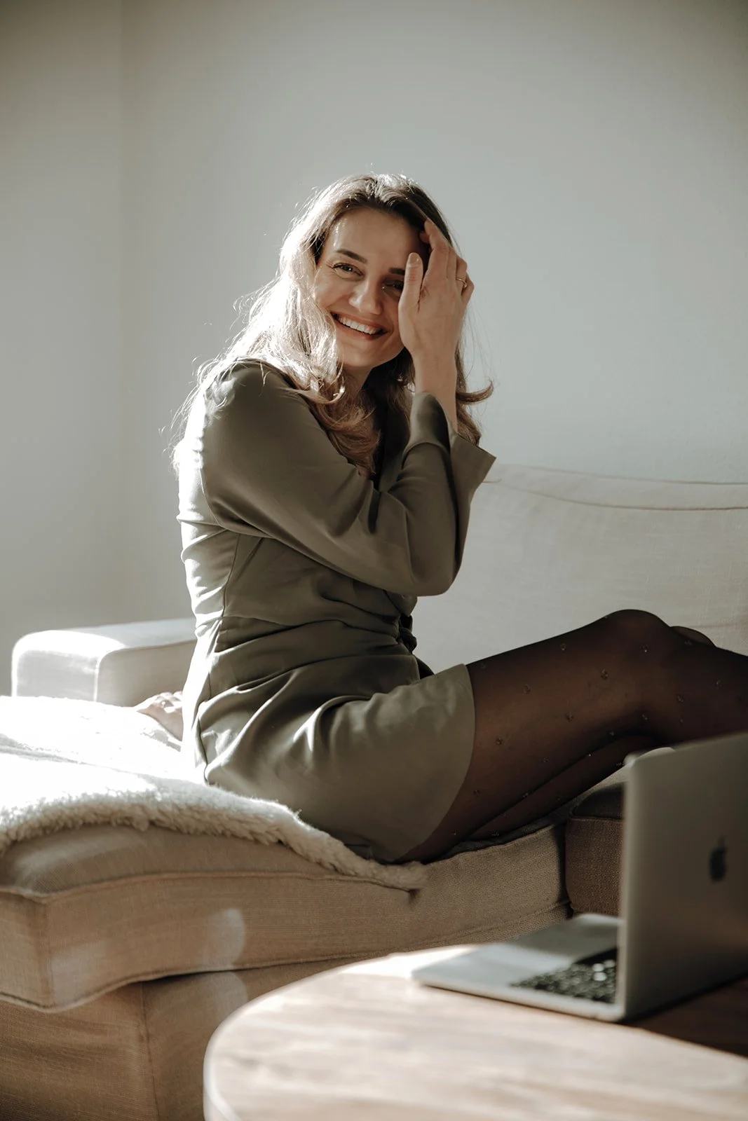 A woman sitting on a beige sofa, smiling, wearing a satin dress and tights, with a laptop on a wooden table nearby.