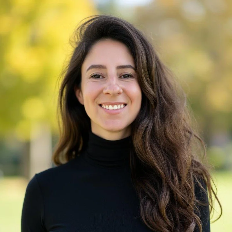 A woman with long, wavy brown hair smiling outdoors during daytime.
