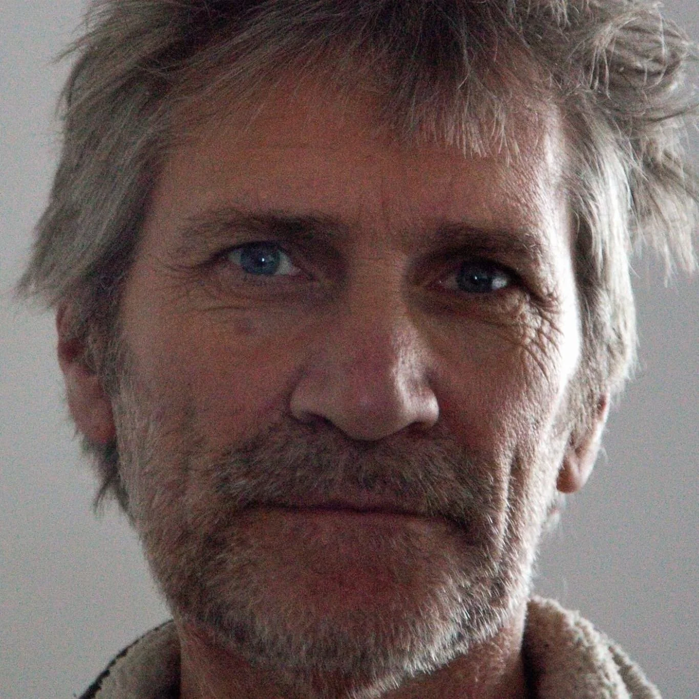 Close-up of an elderly man with gray, tousled hair, blue eyes, and a beard, looking directly at the camera.